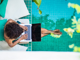 bird view of a remote online working digital nomad women with curly hair and laptop sitting at a sunny turquoise water pool having feet in the water surrounded by cushions and plants in the foreground