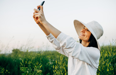 Side view image of a young woman taking a self-portrait on her smart phone on the field background. Female in a white hat and shirt making selfie on a field of flowers on nature background.