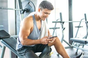 Wall Mural - Handsome young man resting on weight bench and using phone after training at gym.