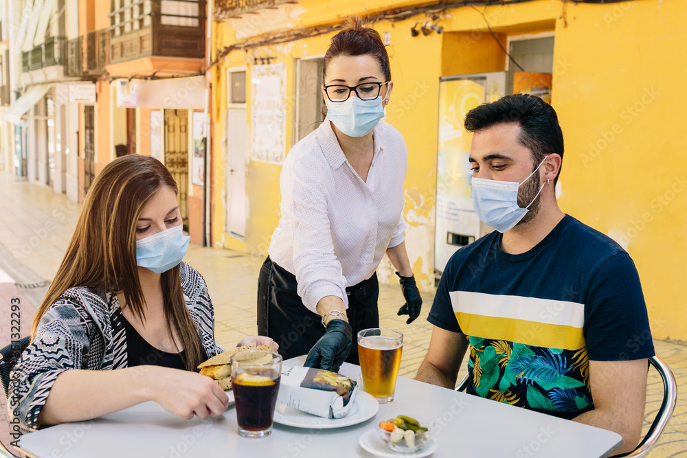 Clients with masks on the terrace of a bar in Spain attended by a waiter with gloves and masks. Social distancing during phase one of de-escalation. Coronavirus - obrazy, fototapety, plakaty 
