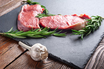 Garlic and raw beef. Two slices of fresh raw meat, garlic and a branch of rosemary on a black slate stone plate. Preparing meat for a barbecue.