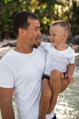 Wall Mural - Happy smile european family have rest and run on white sand beach. Father, long black chestnut hair mother, blond baby boy. white cotton clothes. boho dress. t-shorts.Thailand. Aquamarine crystal sea