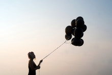 Woman In Silhouette Looking At Black Balloons About To Let Go Set Against A Stark Clean Background And Sky At Sunset
