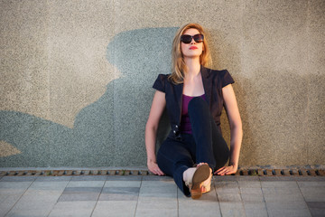 Wall Mural - Sad young fashion woman in sunglasses sitting at the wall on sidewalk