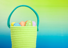 Close Up On A Bright Green Yarn Woven Easter Basket With Light Blue Handle Filled With Festive Colored Eggs With Dots, Graded Background Blue Base To Green Above.