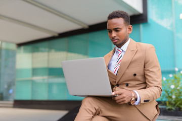 Wall Mural - Young handsome African businessman using laptop while sitting outside the building