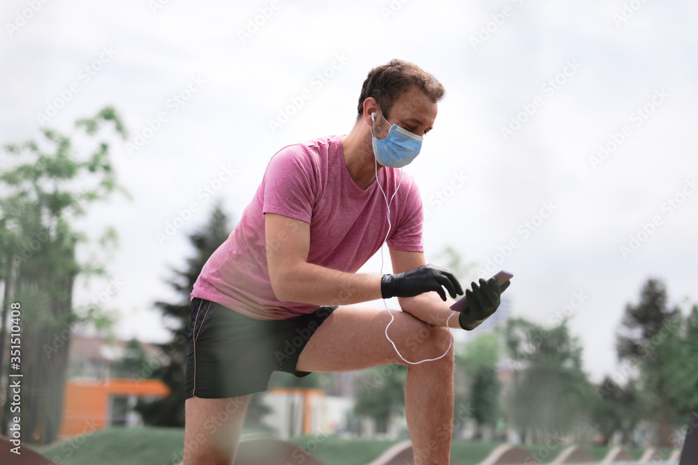 Sportsman with medical mask and gloves, smartphone and earbuds working out, jogging in urban surroundings. - obrazy, fototapety, plakaty 