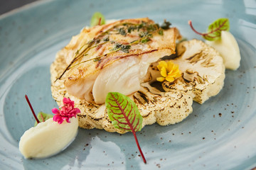 Sea or river baked white fish lies on a base of cauliflower vegetables with sauce. Decoration of flowers and herbs spices. A dish from the chef for serving