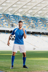 happy professional soccer player in blue and white uniform with ball showing yes gesture on football pitch at stadium