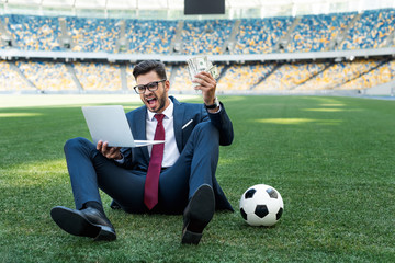 Wall Mural - young businessman in suit with laptop, money and soccer ball sitting on football pitch at stadium and yelling, sports betting concept