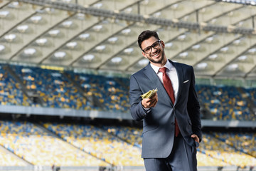 Wall Mural - smiling young businessman in suit and glasses giving money at stadium