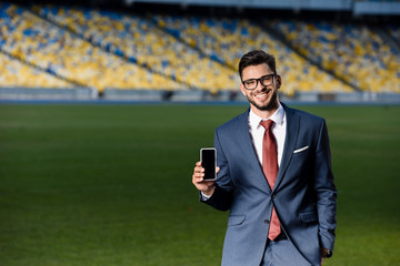 Wall Mural - smiling young businessman in suit and glasses holding smartphone with blank screen at stadium