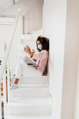 Wall Mural - african american freelancer in medical mask sitting on stairs and showing peace sign while taking selfie near laptop