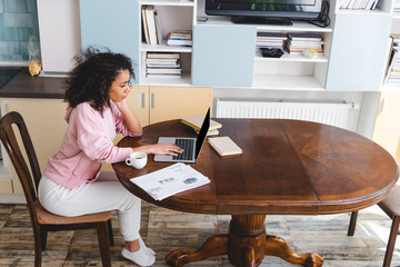 Poster - curly african american freelancer using laptop near cup, books and documents