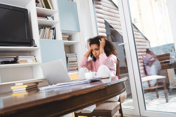 Poster - selective focus of tired african american freelancer looking at laptop near documents, books and cup