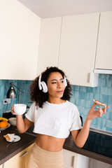 Wall Mural - curly african american girl in wireless headphones holding toast bread with jam and cup of coffee