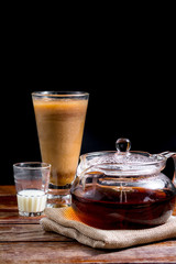 Transparent teapot with hot tea on brown cloth with glass of milk on  wooden table on black background with copy space. Famous beverage of Asian. Healthy drink concept