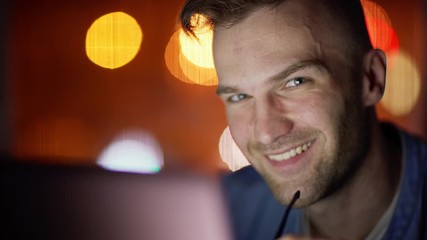 Closeup of focused young man in eyeglasses looking at laptop screen, then taking off eyewear and smiling at camera at night