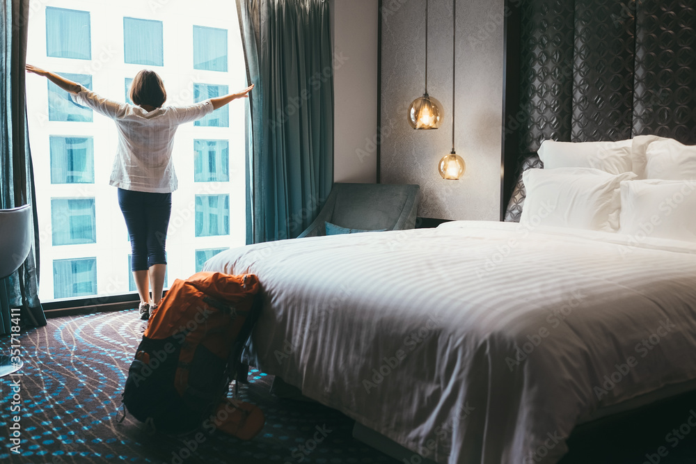 Backpacker female accomodating in 5 stars luxury hotel. She opening a whole wall window curtains to bring a day light into room. Solo traveling with backpack or home returning concept image. - obrazy, fototapety, plakaty 
