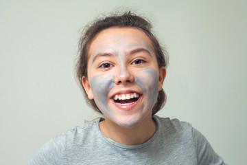 Cute and happy girl smears face with cosmetic clay or mud and smiles. Cosmetic mask, face scrub. The concept of health and beauty.