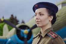 A Young Female Pilot In Uniform Of Soviet Army Pilots During The World War II. Military Shirt With Shoulder Straps Of A Major And A Beret. Against The Background Of A Military Aircraft.