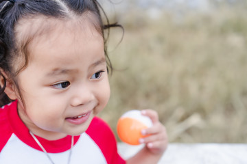 Close up of cheerful and happy adorable little girl smiles with blurred orange and white ball. Cute Asian child female poses outdoor with green summer park background, copy space for text on the right