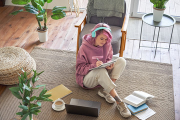 Hipster teen girl school college student pink hair wear headphones writing notes video conferencing on laptop sit in cozy room on floor working learning online in internet watching webinar. Top view.