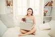 © makistock - Happy beautiful woman working on a laptop and drinking coffee from a mug, while sitting on the bed in the house.