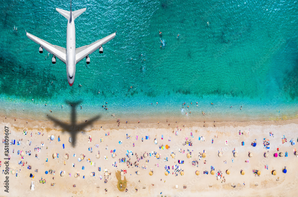 Sommer Reisekonzept mit einem Flugzeug welches über einen vollen Strand mit Menschen und türkisem Meer fliegt - obrazy, fototapety, plakaty 
