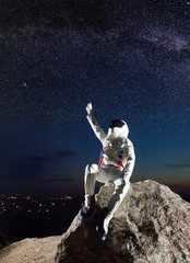 space traveler sitting on top of rocky hill and raising hand while looking at beautiful starry sky. 