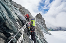 Back View Of Rock Climbers With Backpacks Using Fixed Rope While Ascending High Rocky Mountain. Male Mountaineers Climbing Natural Rock Formation And Trying To Reach Mountaintop.