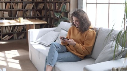 Wall Mural - Smiling young hispanic latina gen z teen girl sit on sofa at home holding phone watching funny social media videos looking at mobile screen, laughing, relaxing in cozy living room using cellphone.