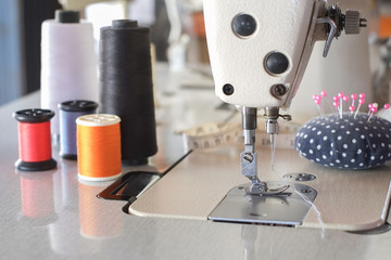 Wall Mural - Needle presser foot of industrial sewing machine for sew cloth and blurred pink pins with pincushion and colorful thread roll with tape measure close-up.