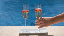 Woman Takes A Glass With Champagne Or Prosecco With Raspberry On Swimming Pool Or Sea Blurred Background. Happy White Woman Put Glass On Poolside In The Sunny Day. Luxury Drink. Romantic Weekend.