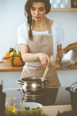 Wall Mural - Young brunette woman cooking soup in kitchen. Housewife holding wooden spoon in her hand. Food and health concept