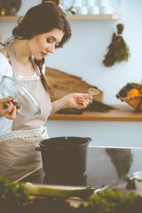 Wall Mural - Young brunette woman cooking soup in kitchen. Housewife holding wooden spoon in her hand. Food and health concept