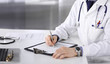 © rogerphoto - Unknown male doctor sitting and working with clipboard of medication history record in clinic at his working place, close-up. Young physician at work. Perfect medical service, medicine concept