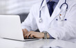 © rogerphoto - Unknown male doctor sitting and working with laptop in clinic at his working place, close-up. Young physician at work. Perfect medical service, medicine concept