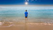 Young boy staring our towards to the horizon with his feet in the surf and a beautiful blue sea and a dramatic sky with a sun star in the distance.