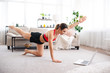 © Admiral - Young woman doing stretching exercise and watching online workout on laptop, fitness training on the floor at home
