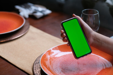 Wall Mural - close up one hand showing green screen smartphone in restaurant. blurred plates on table.