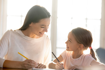 Loving young mother sit at desk studying together with little happy daughter, smiling mom or nanny help teach small girl child, drawing or painting, have fun on weekend at home, education concept