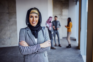 Wall Mural - Successful arab female investor standing in building in construction process with hands crossed and looking at camera. In background is her business partner with agents talking.