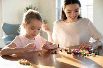 Cute little preschooler girl child and young mom or nanny thread beads making stylish bracelets together, happy caring mother and small daughter engaged in creative weekend activity at home