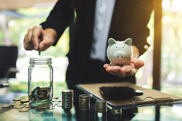 Wall Mural - businesswoman holding a piggy bank while putting coin into a glass jar for saving energy and money c