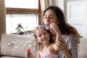 Wall Mural - Close up young mother blowing soap bubbles with smiling little daughter wearing princess dress and diadem, family having fun together, playing funny game, sitting on couch in living room