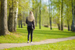 © fotoduets - Young blonde woman slowly walking at town green trees park in warm, sunny spring day. Spending time alone in nature. Peaceful atmosphere. Back view.