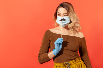 Portrait of positive woman in protective gloves, holding fake mustache accessory over surgical face mask, winking playfully, in good mood in spite coronavirus pandemic threat. studio shot, isolated