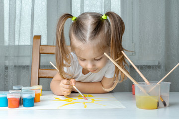 the girl sits on a chair, bent over a drawing and paints the sun with a brush with yellow paint, age 3-4 years