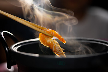 Boiled shrimp in chopsticks holding on hot steam smoke water from hot pot for cooking in seafood party, orange shrimp on black background for people eating to tasty and delicious food in restaurant